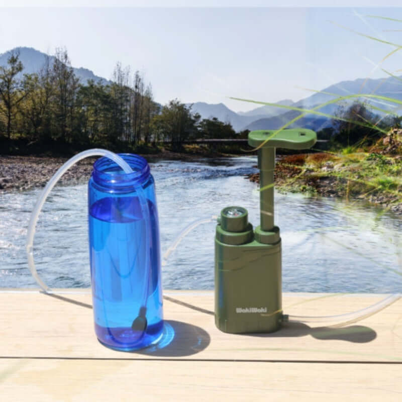 4-Stage Portable Water Purifier Pump next to water bottle by a river with mountains in background.
